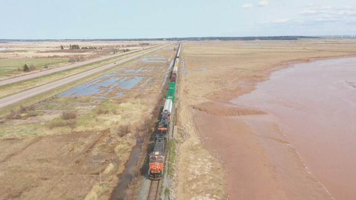 Un train de marchandises est photographié des airs et apparaît de haut en bas au milieu de la photo. Les rails sont très près de la berge et de l'eau.