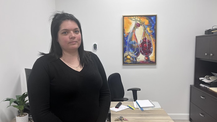 Une femme aux cheveux noirs portant un chandail noir se tient debout dans un bureau. Derrière elle, on voit un tableau coloré accroché au mur blanc, un bureau en bois avec des documents et un meuble de rangement.