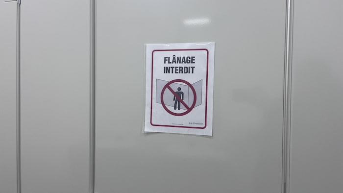 Une affiche blanche fixée sur une surface grise indique « FLÂNAGE INTERDIT » en lettres noires. Un pictogramme rouge montre une silhouette humaine barrée. La mention « La direction » est inscrite en bas à droite de l'affiche plastifiée.