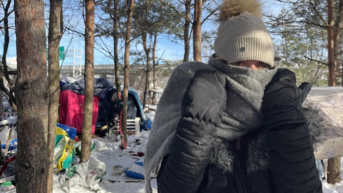 Une personne emmitouflée dans un foulard gris et une tuque blanche se tient devant un campement de fortune en forêt. Le sol est enneigé et jonché de détritus, avec des tentes et une route visibles en arrière-plan.