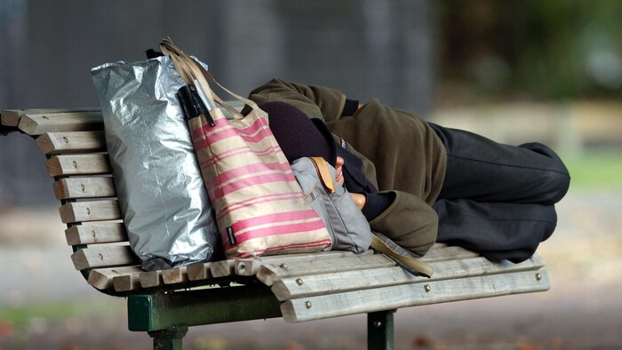 Un homme dort sur un banc de parc, ses effets personnels tenant dans quelques sacs.