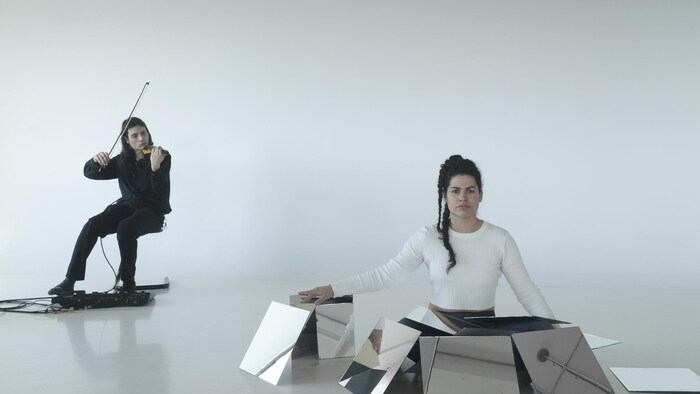 Une femme est entourée de miroirs carrés sur scène, à côté d'un violoniste. 