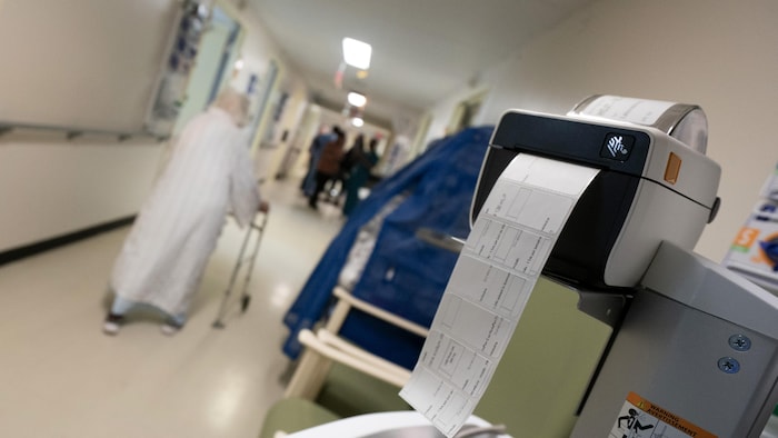 Une machine imprimant des étiquettes à l'hôpital Jean-Talon, à Montréal, avec en arrière-plan une patiente se promenant dans un corridor
