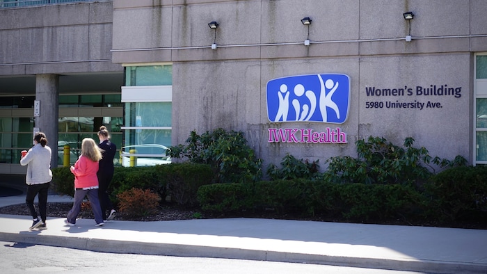 Trois employées de l'hôpital marchent en direction de l'entrée d'un bâtiment du centre IWK, identifié sur sa façade par un logo et par son adresse.