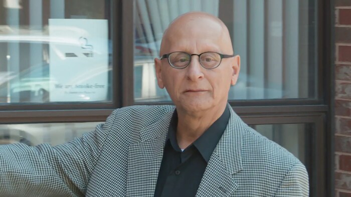 Un professeur chauve portant des lunettes rondes, photographié devant une grande fenêtre aux stores verticaux.