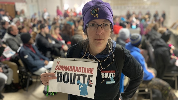 Jacinthe Marcoux est debout dans une salle tenant une pancarte en main, en arrière-plan plusieurs personnes sont assises.    