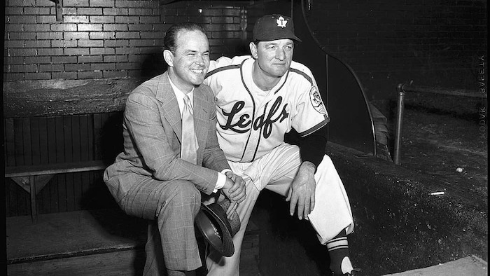 Un homme en complet et un homme en uniforme de baseball sont assis sur un banc. 