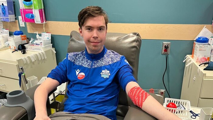 Un homme portant un chandail bleu décoré du logo des Wanderers d'Halifax est allongé dans une chaise longue en cuir. Il a un bandage rouge à l'intérieur du coude après avoir donné du sang.