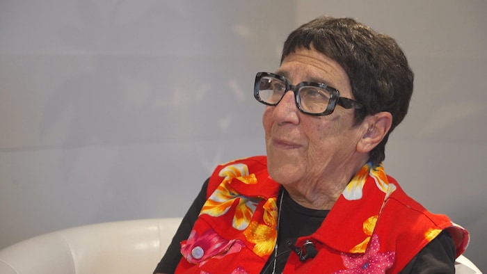 Profil gauche d'une femme aux cheveux courts, portant des lunettes à grosse monture, vêtue d'une criarde blouse fleurie.