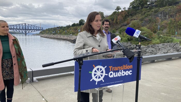 Jackie Smith lors d’une conférence de presse le long de la promenade Samuel-de-Champlain en automne. Elle se tient debout, derrière un lutrin, et est entourée de deux candidats. Au loin, on aperçoit le pont de Québec et le pont Pierre-Laporte.