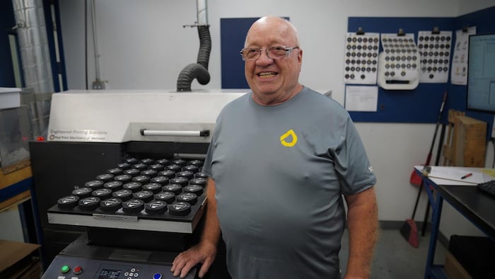 Il est souriant, près d'une machine qui imprime sur des rondelles de hockey.