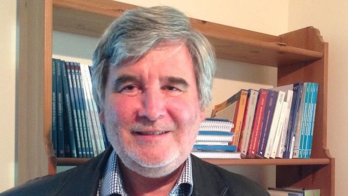 Un homme portant un veston et une chemise, devant une bibliothèque remplie de livres.
