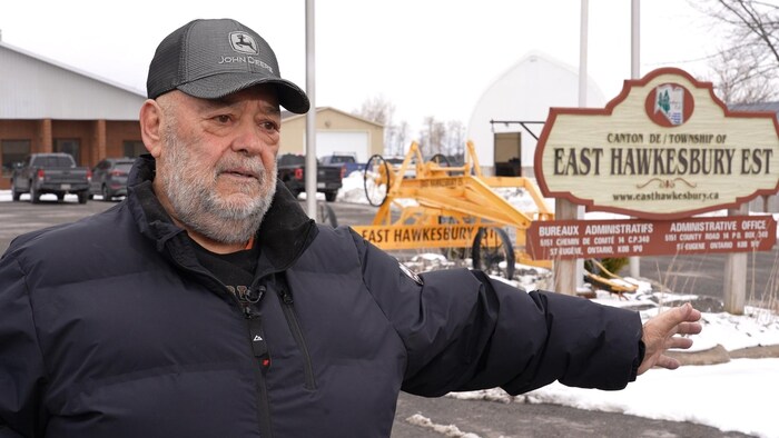 Le maire adjoint Jacques Transmontage porte une casquette et un manteau d'hiver. Il est debout devant l'enseigne du canton de Hawkesbury Est.