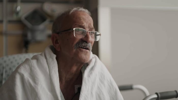Un homme avec des lunettes et une moustache est assis dans une chambre d'hôpital.