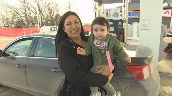 Une femme tient un enfant dans ses bras devant son automobile dans une station-service.