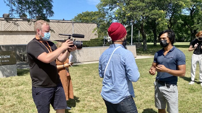 Un homme tient une caméra et filme Jagmeet Singh qui discute avec un autre homme à l'extérieur.