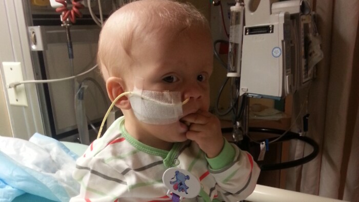 L'enfant est dans un lit d'hôpital, avec un tube de soluté qui se rend à son nez, et a deux doigts dans sa bouche.