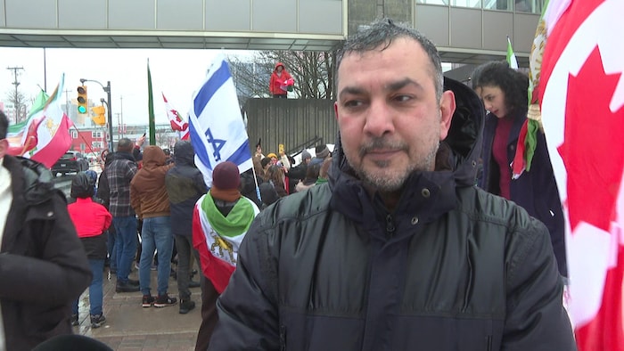Un homme de dos à une manifestation. Des gens agitent des drapeaux de l'Iran, d'Israël et du Canada.