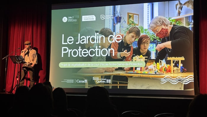 Dans une salle sombre, un écran sur lequel est projetée l'image d'enfants et d'une femme regardant une maquette sur une table.