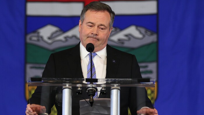 Jason Kenney devant un micro. Derrière lui se trouve un drapeau de l'Alberta. Il a l'air déçu.