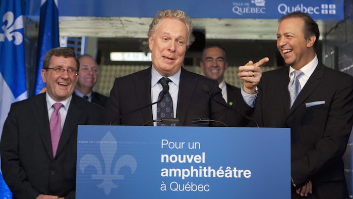 Jean Charest en conférence de presse entouré de Régis Labeaume et Sam Hamad. Une affiche devant le premier ministre porte la mention «Pour un nouvel amphithéâtre à Québec».