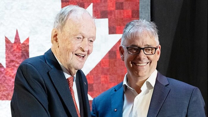 Jean Chrétien, debout devant un mur décoré d'une oeuvre représentant le drapeau du Canada, serre la main de Ronnie LeBlanc.