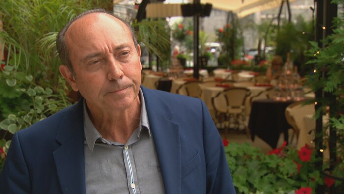 Jean-Claude Crouzet accorde une entrevue à Radio-Canada devant la terrasse extérieure d’un restaurant.