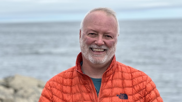 Un homme avec un manteau orange et les cheveux blancs sourit pour la photo.