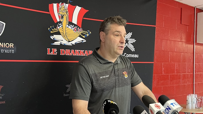 Un entraîneur répond aux questions des journalistes. Derrière lui, un affiche avec l'insigne de l'équipe de hockey du Drakkar.