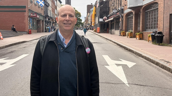 Jean-François Joubert sourit à la caméra, au milieu de la rue Saint-Jean à Québec. La rue est fermée à la circulation automobile et des piétons y marchent, en arrière-plan.