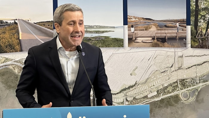 Le ministre parle à un micro derrière un lutrin. Derrière lui se trouvent plusieurs photos et images modélisées de passerelles et divers quais le long du fleuve dans le secteur de Beauport.