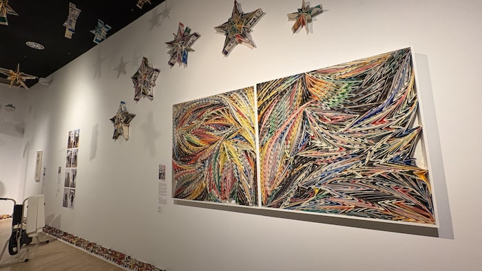 Une grande œuvre colorée faite de pièces de cartons placés sur le côtés pour créer des formes est accrochée sur le mur d'une salle d'exposition. 