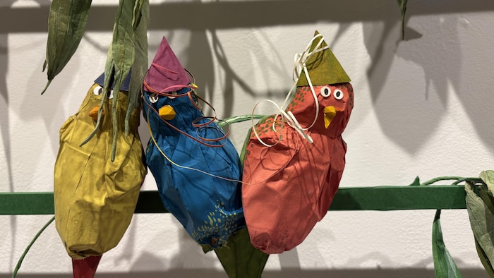 Trois oiseaux en papier coloré et portant des chapeaux coniques sont perchés sur un support vert.
