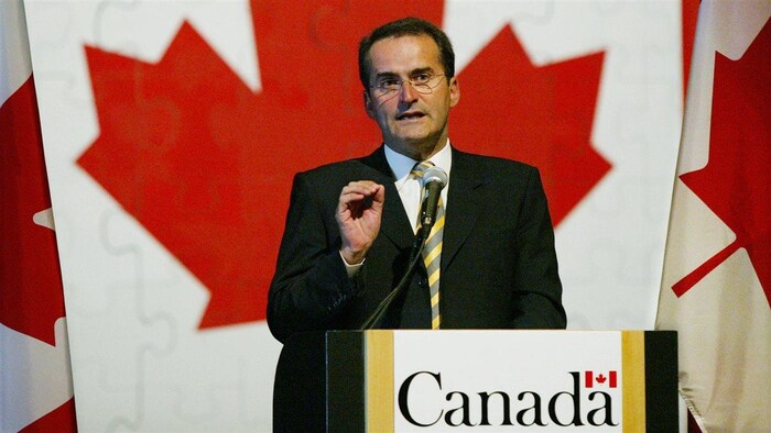 Jean Lapierre s'exprime devant un lutrin. Un immense drapeau du Canada se trouve derrière lui.