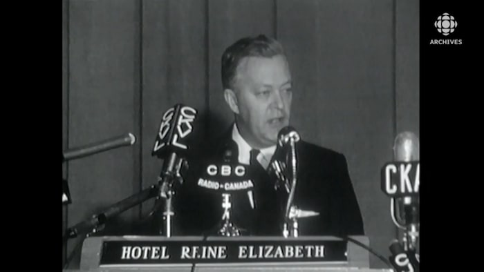 Jean Lesage en conférence de presse, entouré de micros (CKVL, CBC, etc). Inscription « Hôtel Reine Elizabeth » sur le lutrin.  
