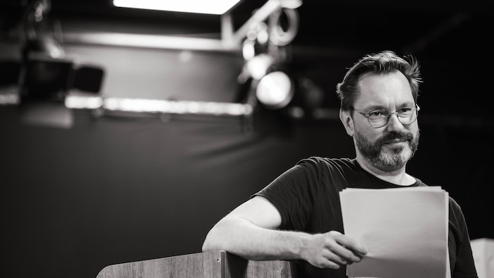 Portrait d'un homme barbu lisant des documents près d'un pupitre sous des éclairages de scène.