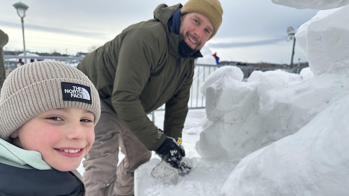 Un père et son fils regardent l'objectif en souriant, alors qu'ils travaillent leur sculpture de neige.