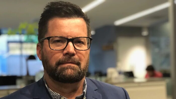 Un homme barbu et portant des lunettes devant des bureaux.