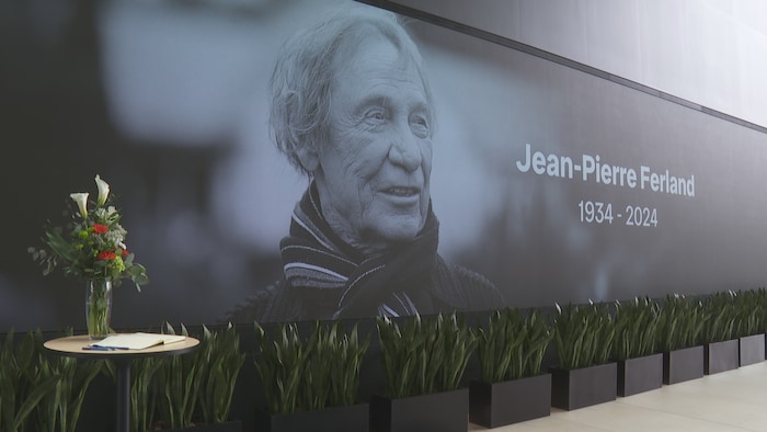 Un bouquet de fleurs et un registre déposés sur une table dans l'atrium de la Maison de Radio-Canada à Montréal devant un immense écran projetant une photo de Jean-Pierre Ferland.