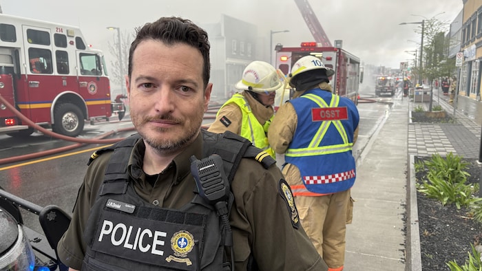 Jean-Raphaël Drolet pose à l'intérieur du périmètre de sécurité pendant la fin de l'incendie. Derrière lui, deux pompiers discutent.