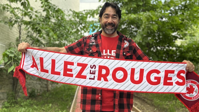 Un homme sourit et tient un foulard sportif où il est inscrit Allez les rouges.