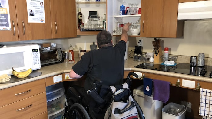 Un homme en fauteuil roulant, de dos, attrape un verre dans une armoire de son appartement, alors qu'il est assis dans son fauteuil roulant.