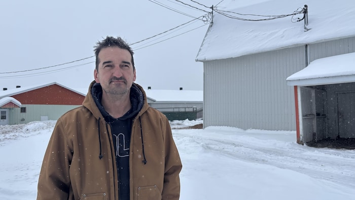 Photo en gros plan d'un homme d'âge moyen portant une veste de travail brune et un capuchon noir, debout à l'extérieur sous une légère neige. Il est positionné devant des bâtiments de ferme recouverts de neige. L'arrière-plan montre des fils électriques suspendus entre les structures.