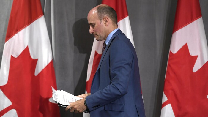 Jean-Yves Duclos, debout et tenant des feuilles, devant des drapeaux du Canada.
