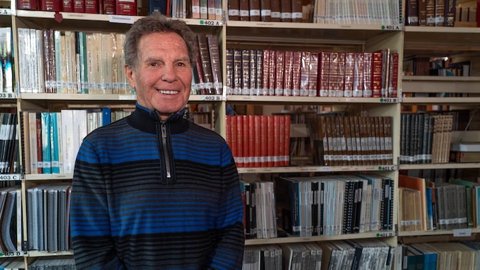 Un retraité souriant, Jean-Yves Vézina, se tient debout devant une étagère de bibliothèque.