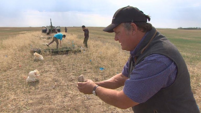 Jeff Schoenau, à droite, tient un petit échantillon blanchâtre de terre compactée, alors que deux personnes examinent le sol à l'arrière-plan.