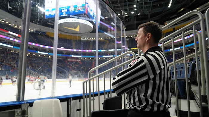 Dans son gilet d'arbitre, il regarde un match au bord de la patinoire