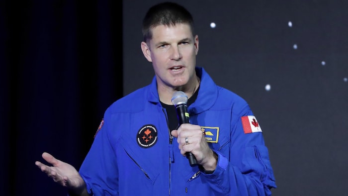رائد الفضاء الكندي جيريمي هانسن يتحدث واقفاً وهو يحمل ميكروفوناً بيده اليسرى.