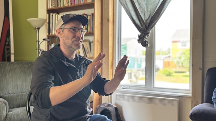 Un homme portant une casquette et des lunettes s'exprime avec ses mains, assis devant une fenêtre et une bibliothèque.