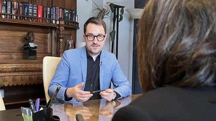Un homme à lunettes et veston bleu discute avec une personne de dos dans un bureau. Derrière lui, une bibliothèque en bois sculpté expose des ouvrages juridiques et un buste décoratif. Il tient un stylo et semble expliquer un sujet.